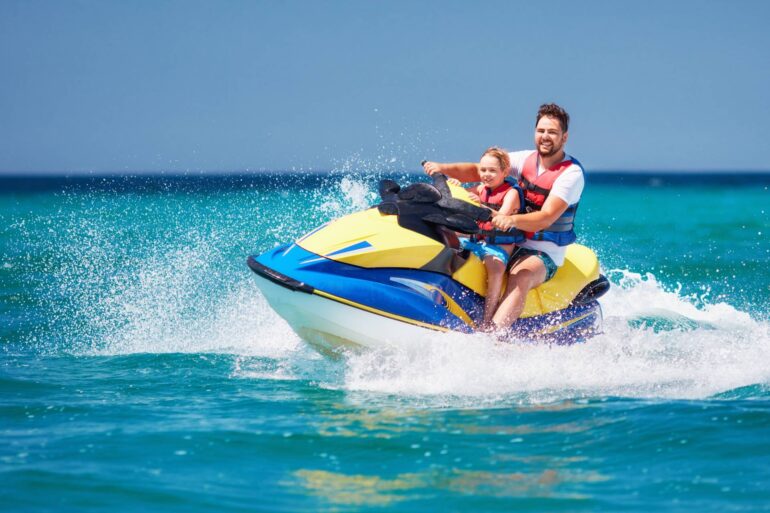 A father and child ride on the back of a jet ski across the water, wearing lifejackets and splashing water in their wake.