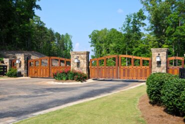 Two wooden gates sit between three brick and stone pillars and block a driveway. Landscaping surrounds the gate.