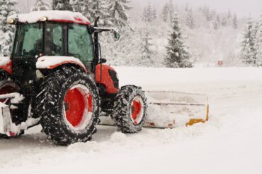 A red and black tractor is moving snow with a yellow plow attachment. Snow covers the ground around the tractor.