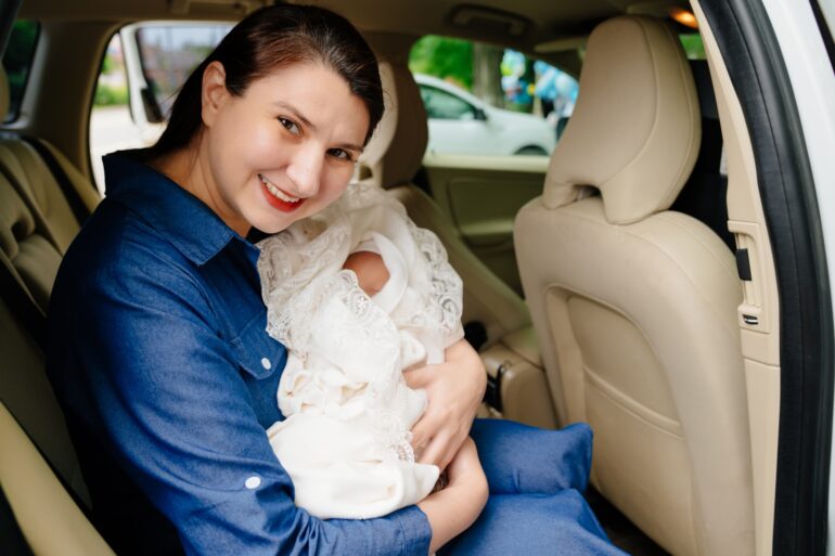 A woman with dark hair, red lipstick, and a denim dress holding a newborn wrapped in white fabric in the backseat of a car.