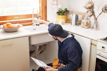 A plumber kneels in front of a kitchen sink with the cabinet door open. He is reaching up to adjust the faucet.