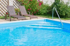 Two reclining lounge chairs lie at the edge of a pool. A rose bush and a white brick building are in the background.