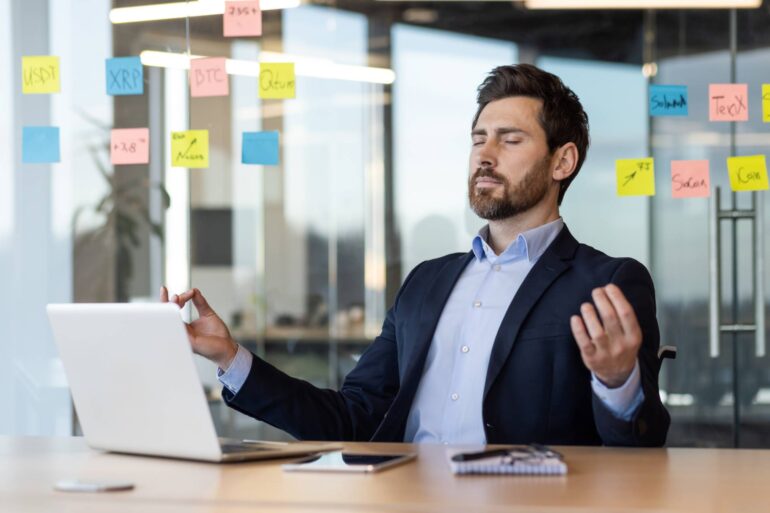 A man sits in a chair at an office with a desk and laptop in front of them while, with eyes closed, they try to meditate.