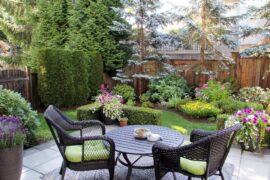 A small backyard filled with trees, bushes, and flowers surrounded by a wooden fence. There is a patio with a table and chairs.