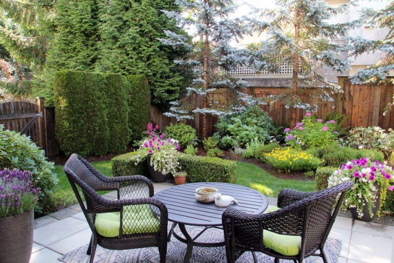 A small backyard filled with trees, bushes, and flowers surrounded by a wooden fence. There is a patio with a table and chairs.