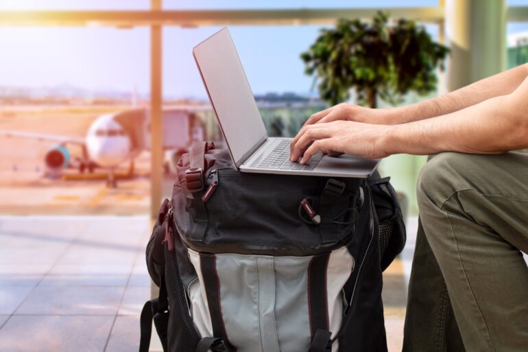 A man sitting in an airport, using his large backpack as a desk to type on his computer. A plane is parked for boarding outside.