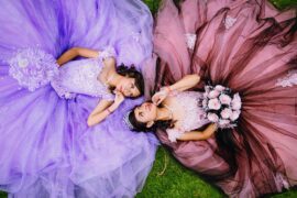 A teenage girl in a lilac ballgown lays on the grass across from a teenage girl in a dusty rose ballgown; both hold bouquets.