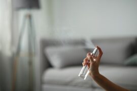 A close-up view shows a hand holding a clear glass bottle while the finger presses on the top, releasing a mist.