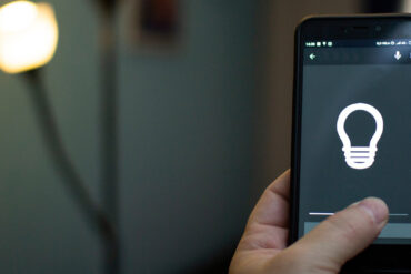 A close-up view shows a hand holding a phone with the thumb pressing a gray button on the screen. A lightbulb is on the screen.