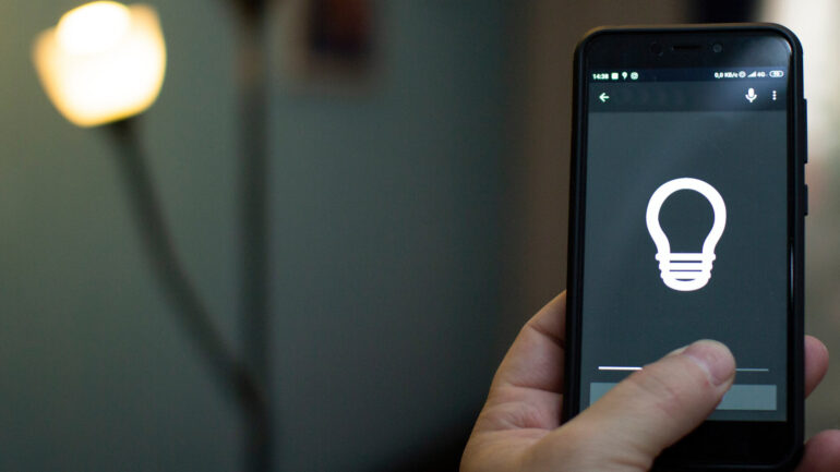 A close-up view shows a hand holding a phone with the thumb pressing a gray button on the screen. A lightbulb is on the screen.