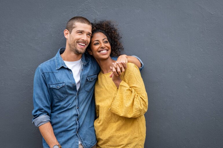 A close-up of a young couple in love smiling, standing, and holding hands while looking off to the right.