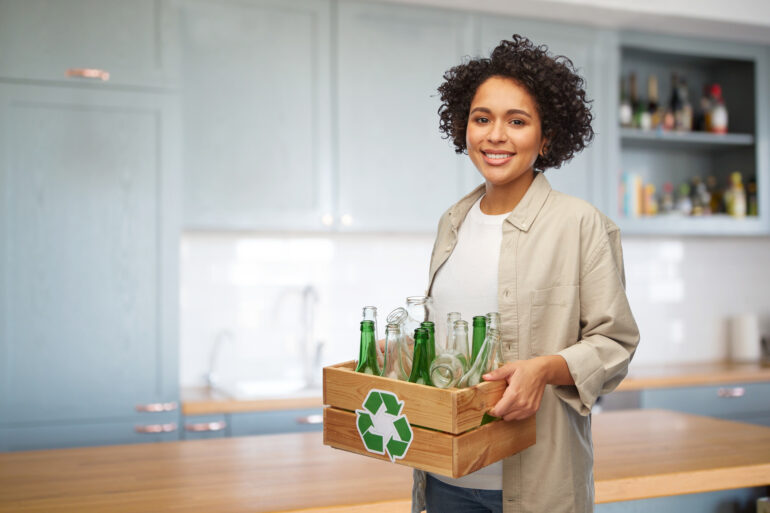 A woman smiles and holds a wooden box with empty glass bottles. The box also has a recycling sign on it.