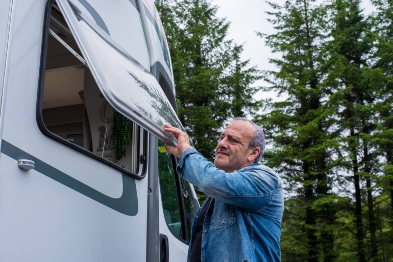 A man wearing a jean jacket stands outside while closing a window to an RV. Tall trees appear in the background.