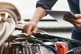 An automotive mechanic standing at the front of a car with the car's hood open. They have their hand on the engine.