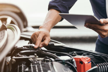 An automotive mechanic standing at the front of a car with the car's hood open. They have their hand on the engine.