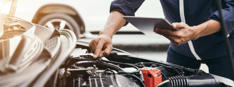 An automotive mechanic standing at the front of a car with the car's hood open. They have their hand on the engine.