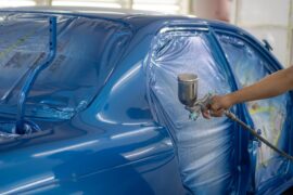 A spray painting gun is held up to a car that is in the process of being painted a bright shade of blue.