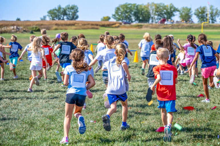 Children running around in a field in athletic wear on a sunny day with cones on the ground in the grass.