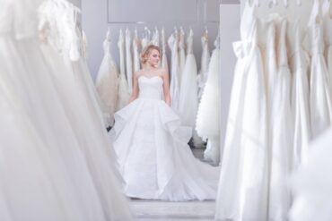 A woman in a wedding dress standing amidst a collection of wedding gowns, all hanging neatly on metal racks along the wall.