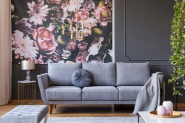 A gray couch sitting in front of a wallpapered wall that has pink florals on it. There's a hanging light fixture overhead.