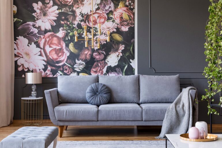 A gray couch sitting in front of a wallpapered wall that has pink florals on it. There's a hanging light fixture overhead.
