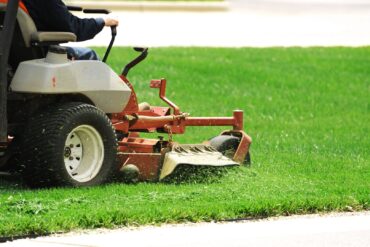 A riding lawnmower is driven across a verdant lawn, cutting the grass and shooting it out to the side.