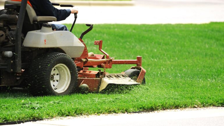 A riding lawnmower is driven across a verdant lawn, cutting the grass and shooting it out to the side.