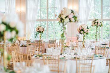 A large room contains several tables covered in white tablecloths, glass cups, tall floral bouquets, and gold chairs.