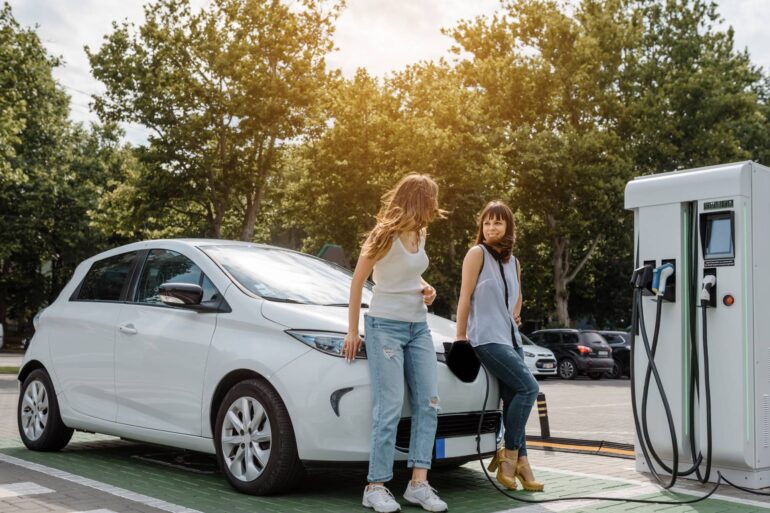Two young women lean against the hood of a white EV charging at a public charging station. Trees line the background.