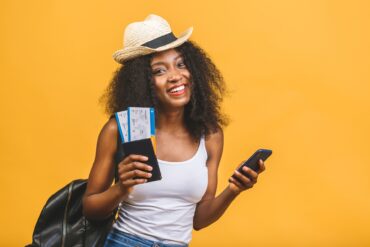 Against a solid yellow background, a young smiling woman holds her passport and tickets in one hand and her phone in the other.