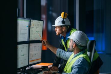 Two engineers wearing protective gear and helmets looking at multiple computer screens displaying information.