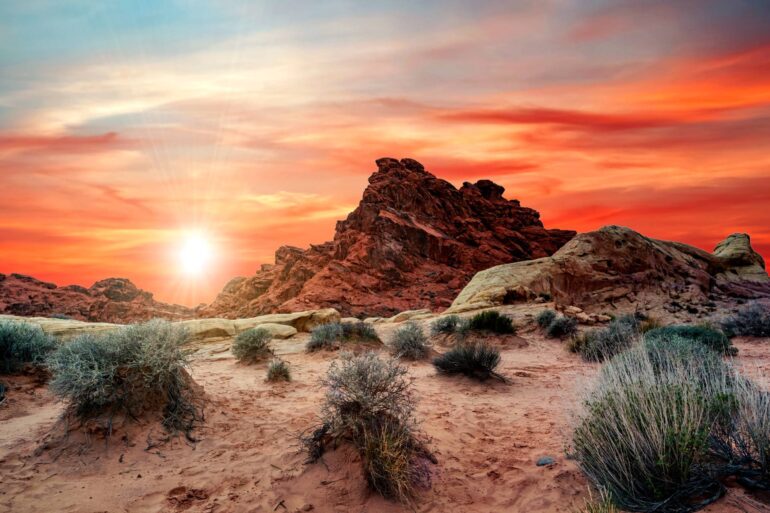 A large red rock formation stands tall in the desert, with small bushes at the base. The sunset turns the sky orange and red.