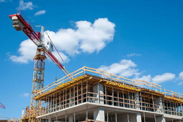 A multi-story building under construction on a sunny day. A large red crane is towering above the structure.