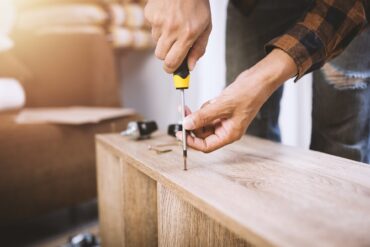 An extreme close-up of a person using a yellow screwdriver to put a small screw into a piece of wooden furniture.