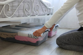 Someone's arms and legs, up close, as they put two plastic storage bins of clothes underneath a bed.