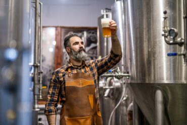 A man wearing a brown leather apron holds a glass full of an amber liquid up in the air and looks at it.