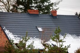A red brick house with a brick chimney and large black shingle metal roof where white snow melts and falls to the edge.