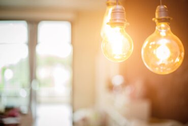 In the foreground, two bulbs with yellow light glow. In the background, sunlight shines through a window or glass door.