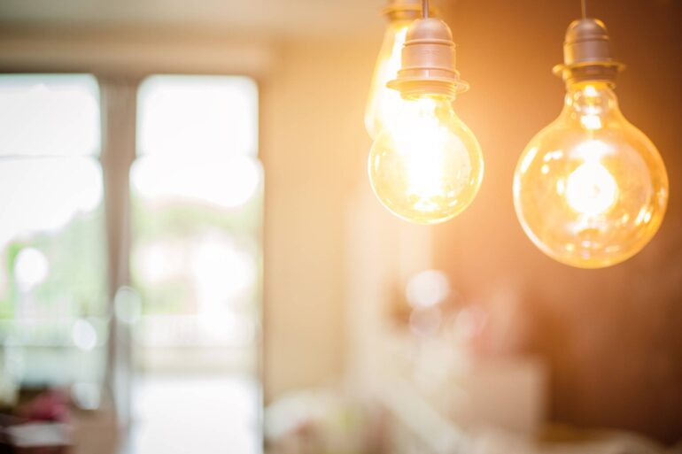 In the foreground, two bulbs with yellow light glow. In the background, sunlight shines through a window or glass door.