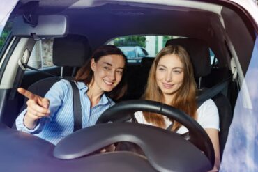A mother sits in the passenger seat of a car and teaches her teenage daughter how to drive. Both are smiling.