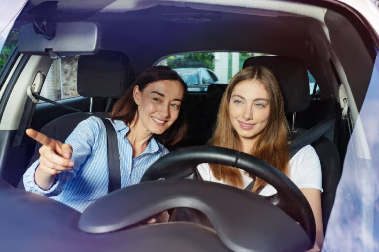 A mother sits in the passenger seat of a car and teaches her teenage daughter how to drive. Both are smiling.