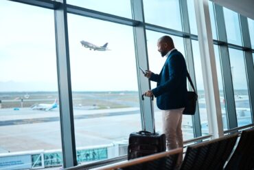 A person in a business suit has a messenger bag and suitcase with him while he checks his phone at the airport.