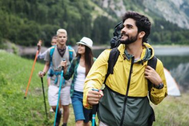 Four people are hiking through the wilderness. Behind them there are lots of trees, a lake, and a mountain.