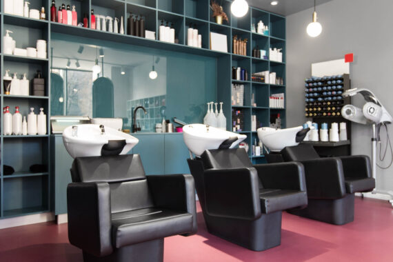 A salon with three hair washing stations. The chairs look quite comfortable and there is a large shelving unit behind them.