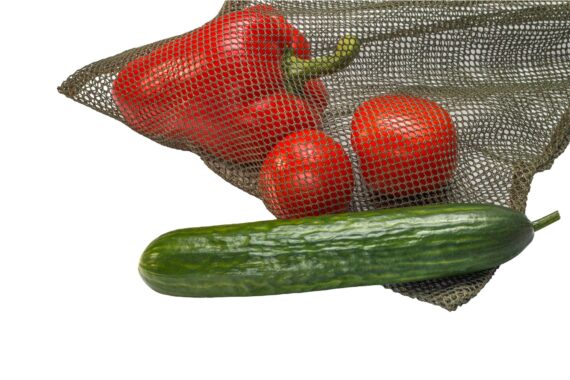 A green mesh laundry bag being used for food. There's a red pepper and tomatoes inside the bag and a cucumber outside of it.