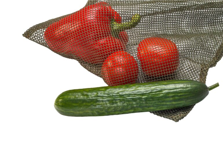 A green mesh laundry bag being used for food. There's a red pepper and tomatoes inside the bag and a cucumber outside of it.