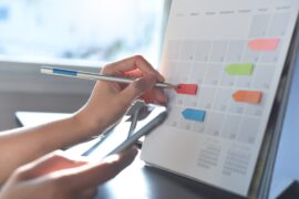 A person places colored sticky notes on a large calendar. They write on the notes and read from their phone.