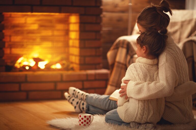 A woman and her child sit on a rug and wear matching sweaters while they curl up with a book in front of the fireplace.