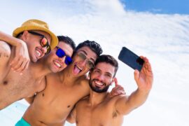 Four men in swimsuits stand on a beach. They huddle together with their arms around each other to take a selfie.