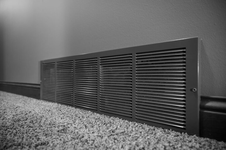 A gray residential vent sits on a gray wall that has gray trim. The flooring is a gray, fuzzy carpet.